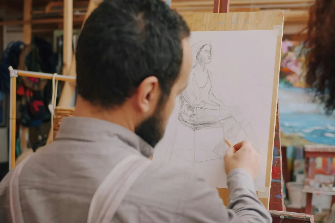 Sesión de dibujo del natural en un taller: una lámina sujeta al caballete con un apunte a grafito de una figura sentada.